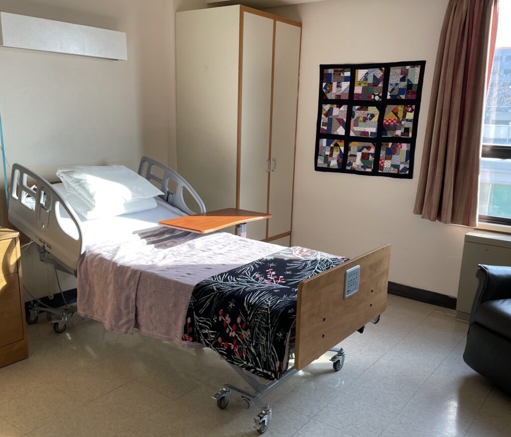 Sample care suite at Dorothy Ley Hospice's temporary location in the McCall Centre. Pictured is a sun filled room with a large window, hospital bed, colourful quilt on the wall and a reclining chair.