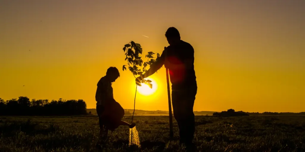 Silhouette of a parent and child planting a tree with a bright sun in the background