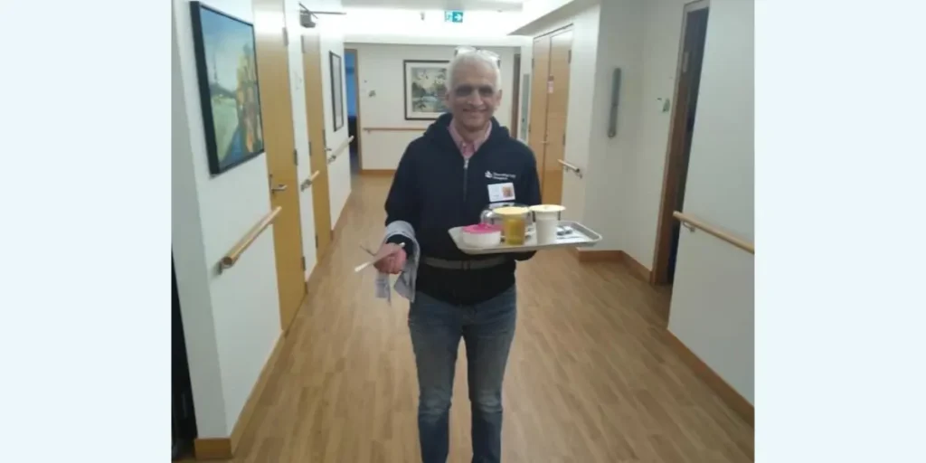 A picture of Dorothy Ley Hospice volunteer Errol, smiling, inside the hospice, carrying a tray of food and beverages.
